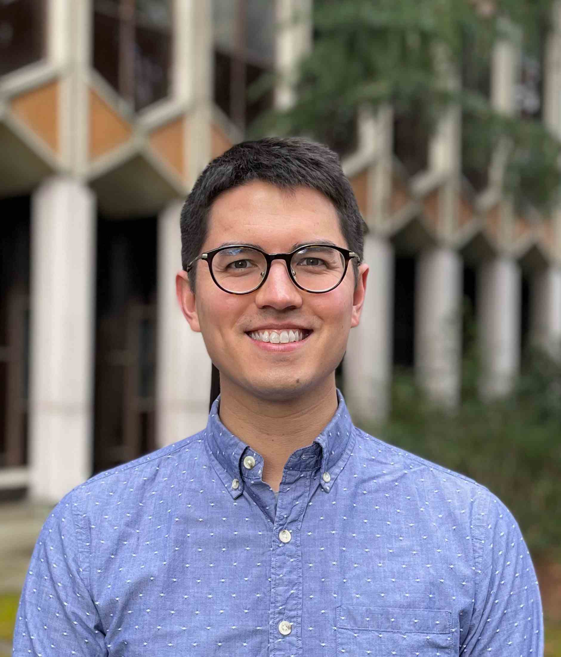 A professional portrait of Douglas Reed, a person with short black hair, light tan skin and brown eyes. He is wearing round black rim glasses and a periwinkle blue collared shirt.